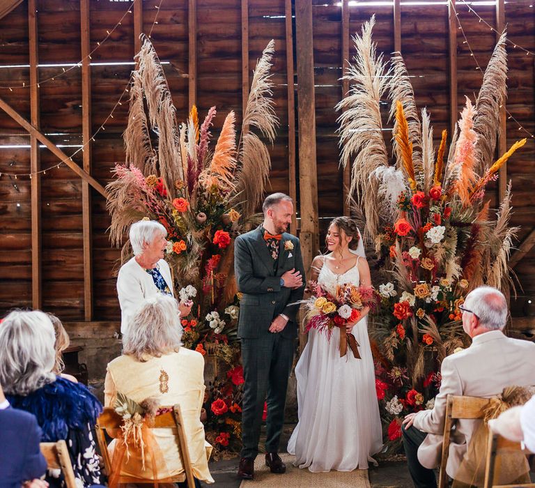 Smiling bride in thin strap wedding dress holding mixed rustic bouquet stands with groom in three piece green suit and orange bow tie by rustic pampas grass installations during barn wedding ceremony