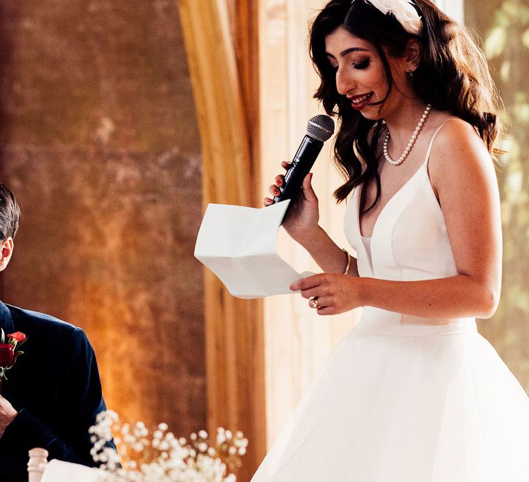 Bride in layered tulle skirt wedding dress and pearl necklace holding microphone makes speech at wedding reception