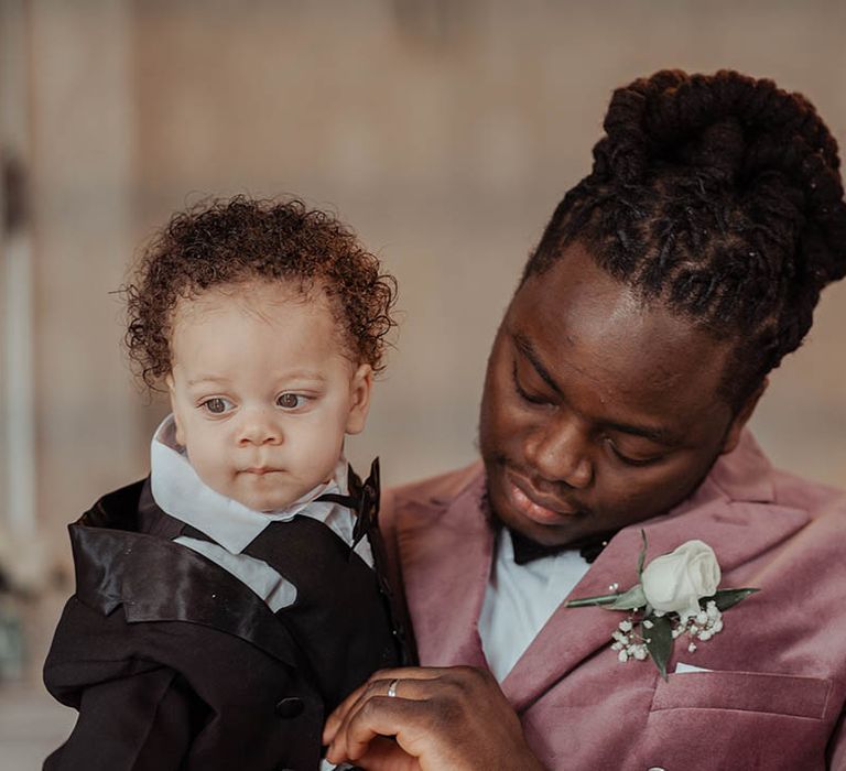 Groom holds his son on his wedding day who wears suit with black tie 