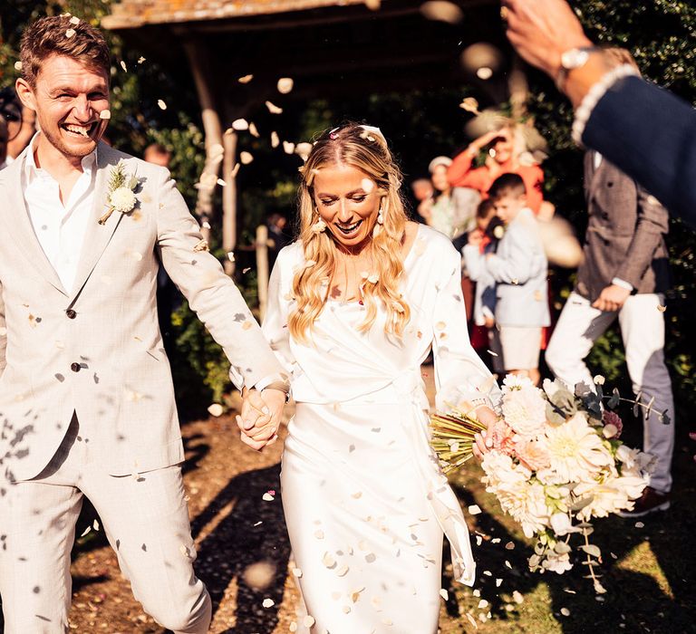 Smiling bride in satin dress and long sleeve wrap top holding mixed bridal bouquet holds hands with groom in open white shirt and grey suit as guests throw petal confetti 