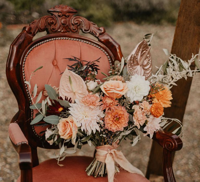 pink, peach, coral and orange wedding bouquet with roses, dahlias, anthurium and foliage resting on a chair 