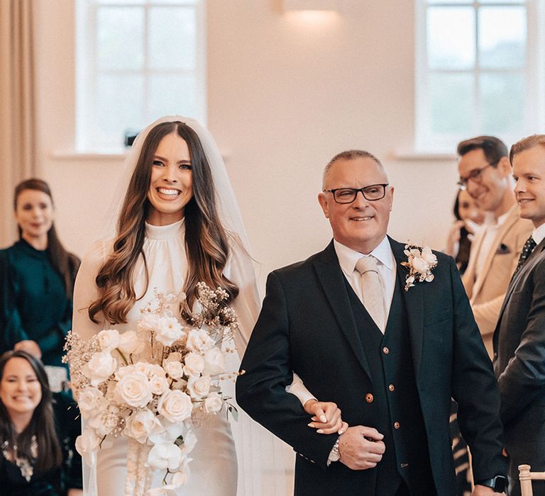 Father of the bride walking his daughter down the aisle in a long sleeve wedding dress as she carries an all-white wedding bouquet 