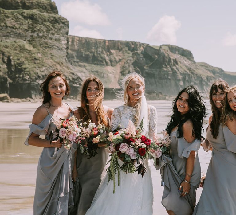 Bride in long sleeve wedding dress stands on beach with bridesmaids in blue open shoulder bridesmaid dresses