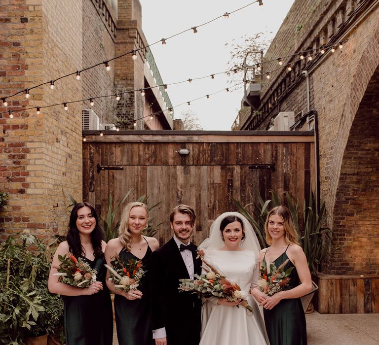 Bride and groom pose with bridesmaids in satin forest green gowns from Rewritten with matching autumnal wedding bouquets