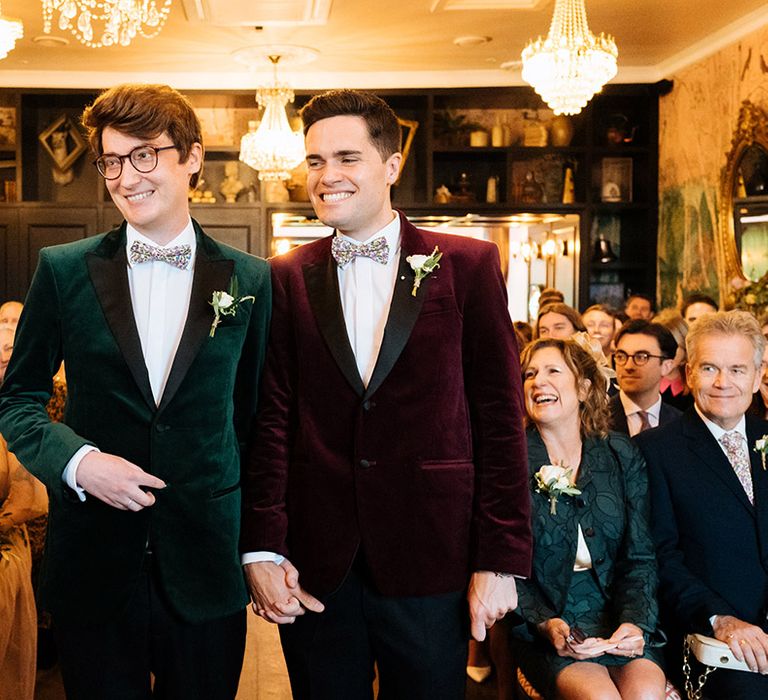 Grooms in velvet tuxedo jackets walk down the aisle together at fun wedding venue with hanging crystal chandeliers