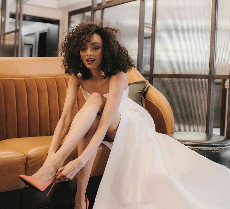 Black bride in a. strapless wedding dress with naturally curly hair, red lipstick and dangly earrings putting on her orange stiletto wedding shoes 