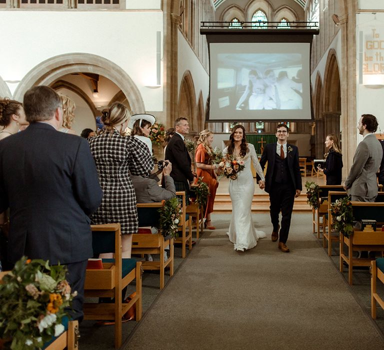 Bride and groom walk back down the aisle as a married couple with a projector above them featuring their best couple pictures