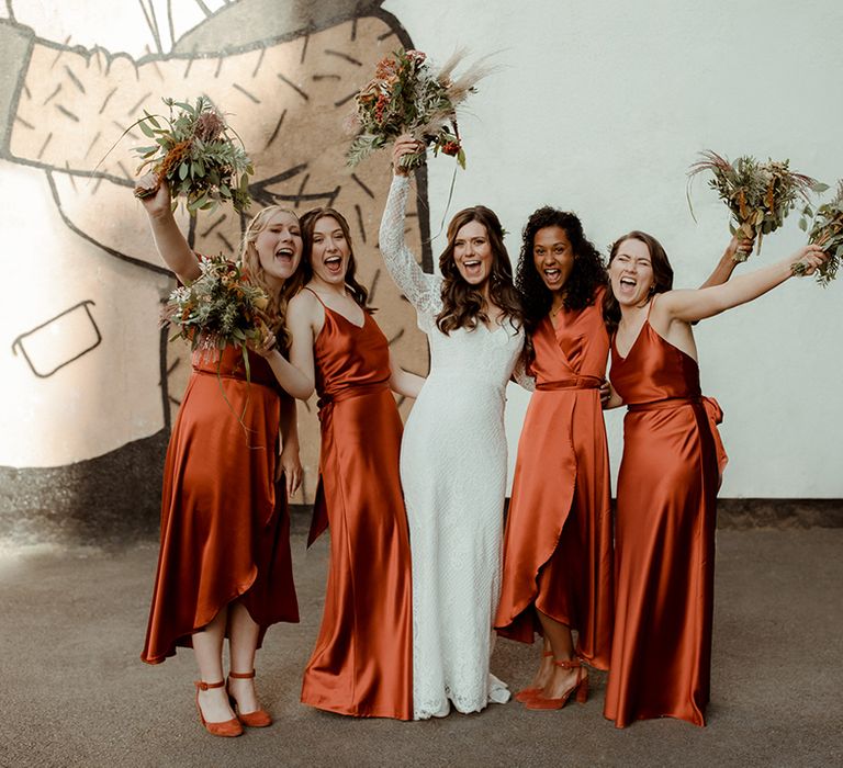 Bridal party lift up their autumnal bouquets with bride in lace Maggie Sottero wedding dress and bridesmaids in orange satin dresses