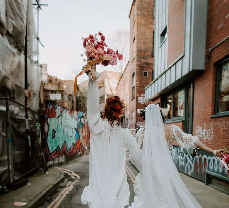 Bride in a short wedding dress holding her red and pink bouquet in the air arm in arm with her bride in a long pearl embellished wedding veil 