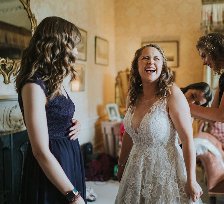 Bridesmaids in deep purple dresses help the bride into her wedding dress 