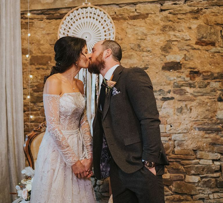 Bride in grey wedding dress and groom in dark grey suit share a kiss in front of macrame wedding decor