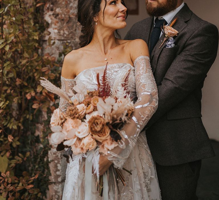 Bride and groom look into eavh other's eyes as the groom hugs the bride from behind wearing a dark grey suit and dark blue tie 