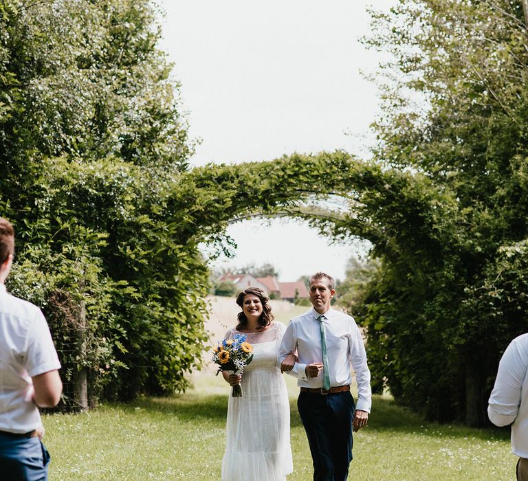 Bride walked down the aisle by the father of the bride in white shirt and green tie with dark trousers for outdoor ceremony 