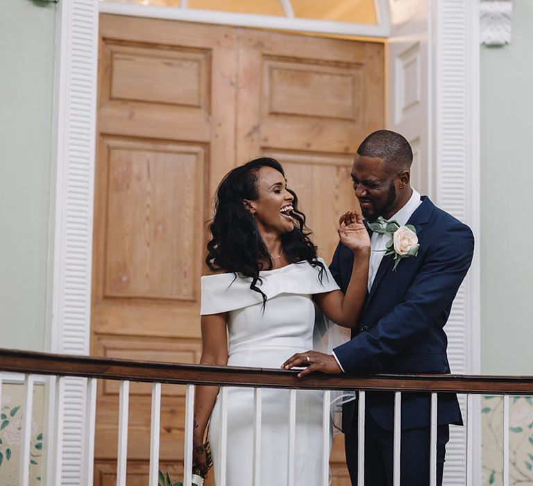 Laughing bride in off the shoulder wedding dress reaches up to touch the groom's face in blue suit and mint green bow tie