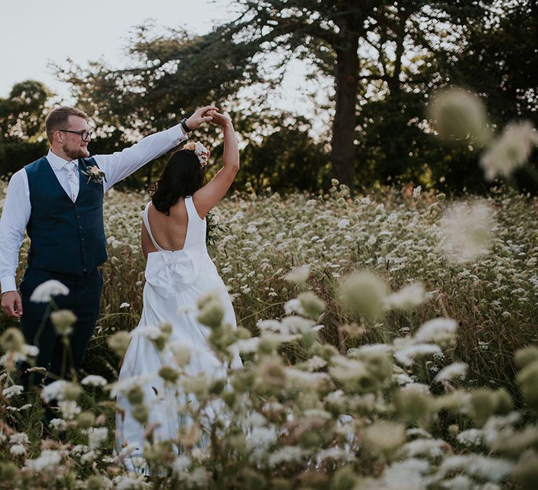 Groom in blue waistcoat spins the bride around wearing white wedding dress with custom bow on the back in the grounds of St Tewdrics House