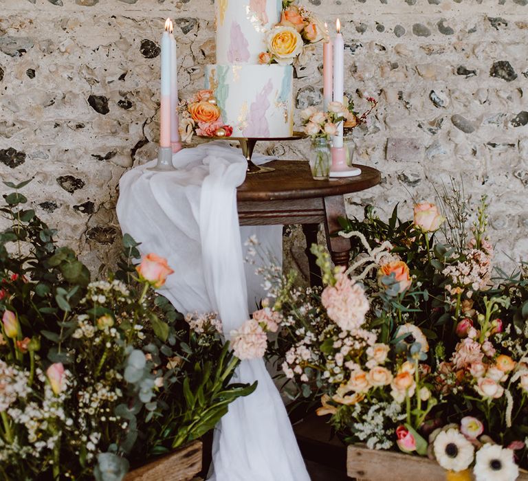 Three-tier white buttercream wedding cake with pastel pink, yellow, green and blue and lilac design surrounded by ombre candles and wooden crates 