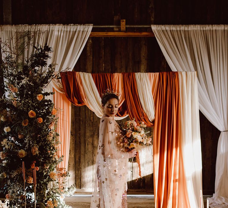 Bride in a gold star celestial wedding dress standing in front of an orange ombre draped altar at Montague Farm Hankham