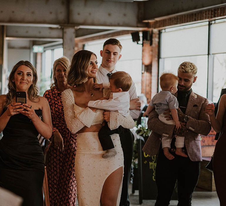 Bride in white pearl co ord holding son with wedding guests 