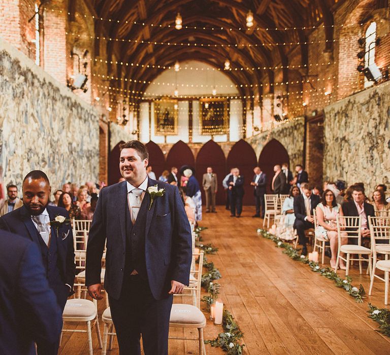 Groom and groomsmen stand together at the front of the ceremony room in matching blue suits with pale grey ties 