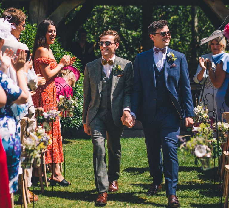 Outdoor wedding ceremony with two grooms walking down the aisle hand in hand wearing sunglasses 