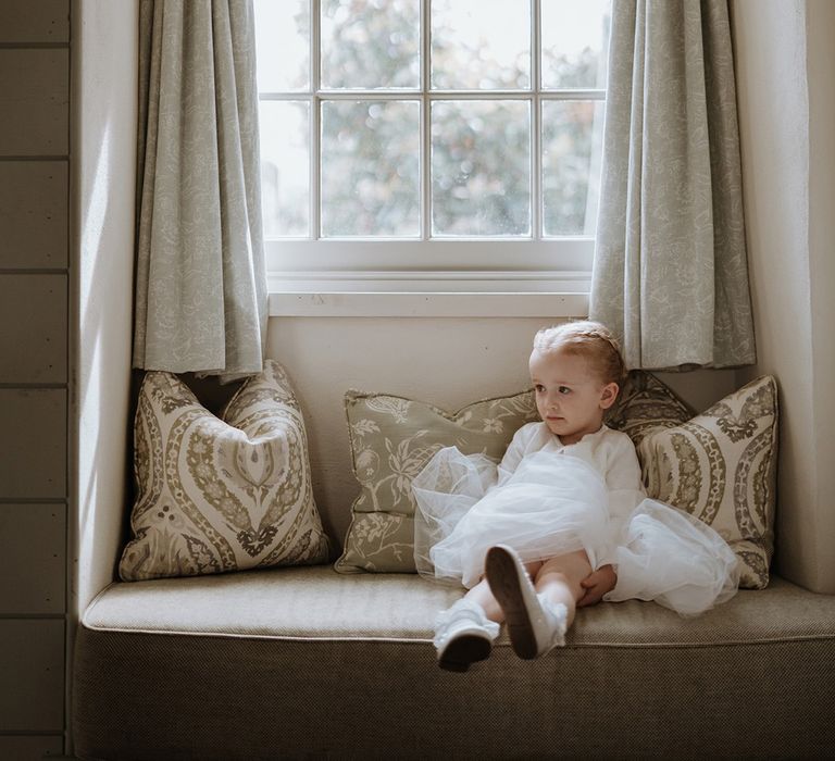 Bride and groom's daughter sits on a window seat in a white tulle dress 