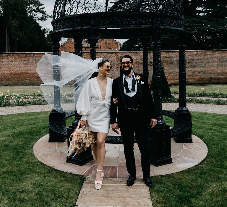 Groom in a tuxedo with his bride in a short wedding dress, sunglasses, platform shoes and bow veil in the gardens at Garthmyl Hall