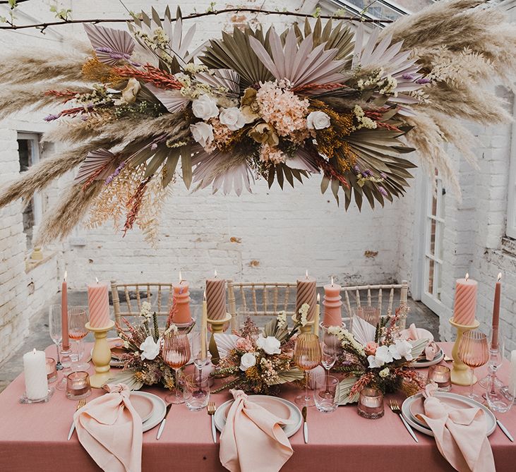 Wedding reception table deco with a pink table cloth, pillar candles mixed with cream candlesticks and preserved pastel flowers and dried grasses 