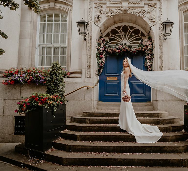 Bride outside their wedding ceremony venue in simple wedding dress with beaded top detail and veil blowing in the wind 