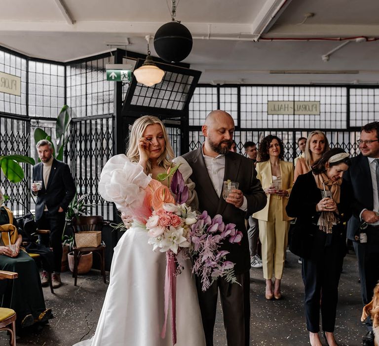 Bride in a JESUS PEIRO wedding dress holding a pastel wedding bouquet crying with her husband in a doubled breasted jacket during the drinks reception speeches 