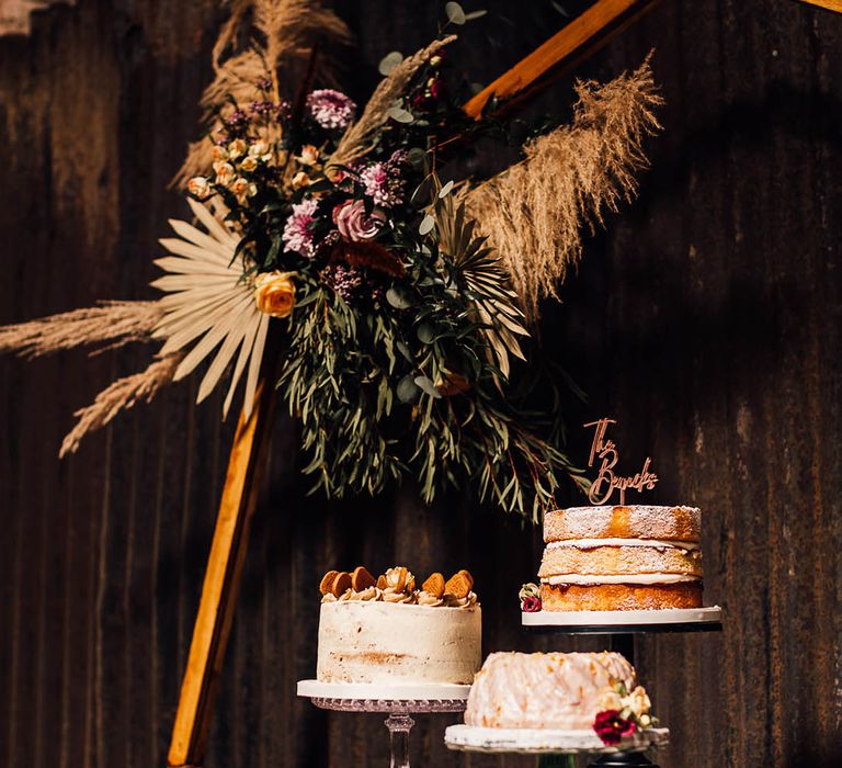 Three wedding cakes on mismatched cake stands next to a hexagonal shape moongate decorated with dried boho flowers 