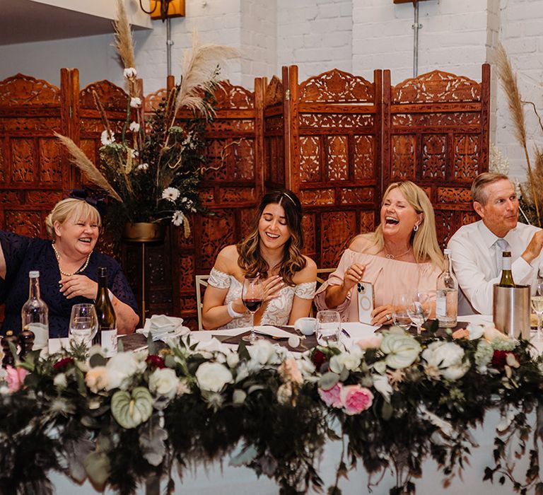 Bride sits with wedding guests at the wedding reception with a boho and rustic style with the wooden screen and dried flowers 