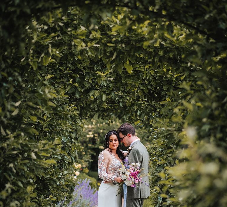 Bride wearing lace long sleeve wedding dress & groom in three piece suit stand within gardens outdoors at the West Green House 