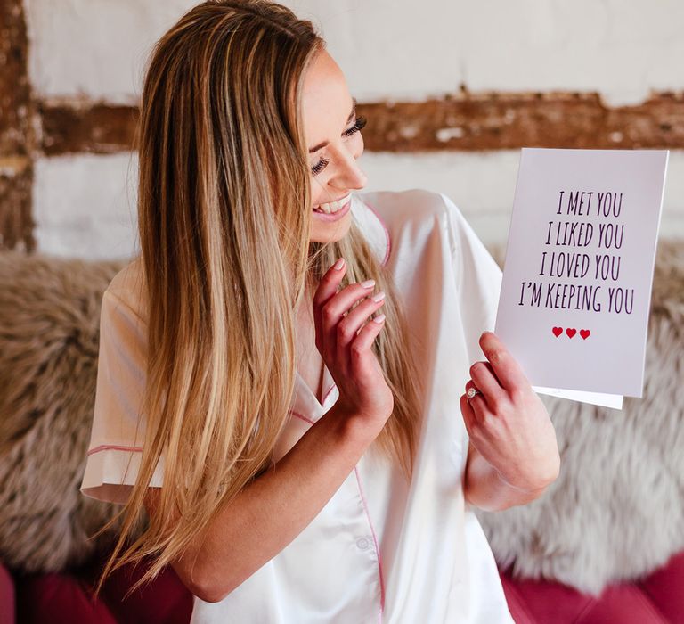 Bride with long blonde hair in white satin pyjamas opens a card from the groom on the morning of the wedding 