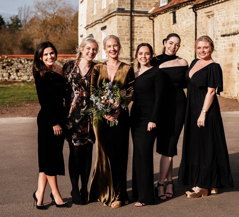 Bride wearing olive green velvet wedding dress stands with her bridesmaids who wear black bridesmaids dresses in different styles 