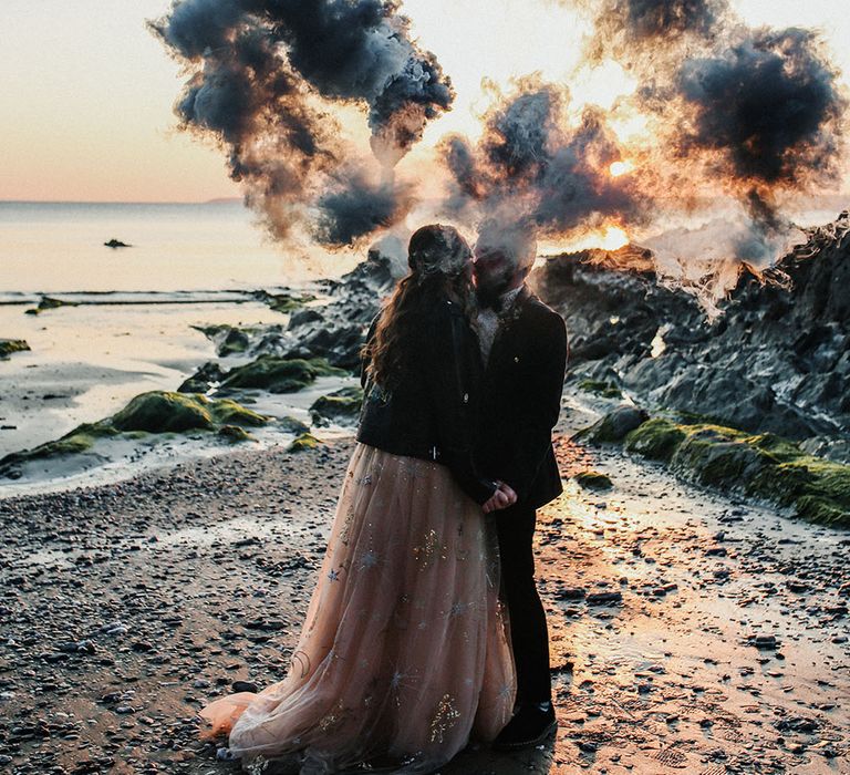 Bride & groom hold colourful smoke bombs on their wedding day along the beachfront in Cornwall 