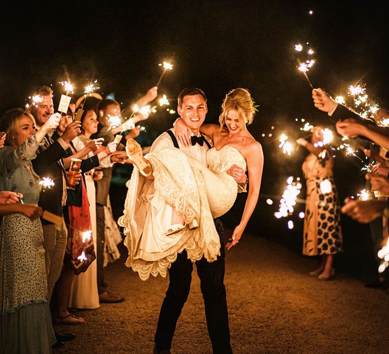 Groom wearing a black bow tie carries the bride in between their guests as they wave sparklers