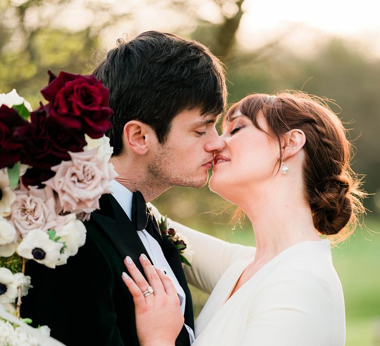 Bride wears 1/4 sleeve wedding dress and her hair in low-bun as she kisses her groom in black tie 