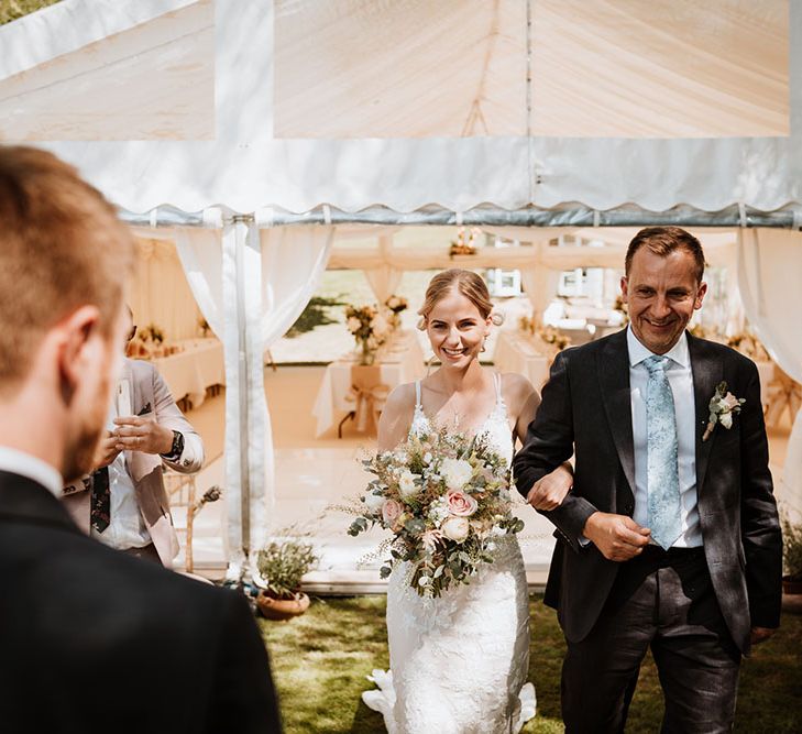 Bride holds pastel floral bouquet and walks down the aisle with her father in front of marquee for outdoor wedding ceremony