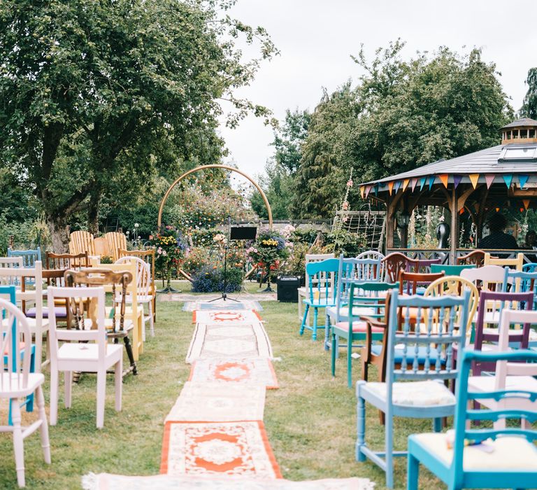 DIY outdoor back garden wedding with Moroccan styled rugs multi-coloured chairs lining the aisle and pom pom wedding decorations