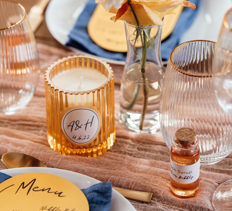 Flower stems, personalised candle and fluted glasssware with blue napkins, yellow menus and a pale pink runner 