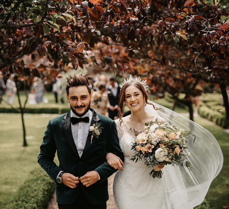 Groom in a green velvet custom suit jacket walks with the bride in a dotted wedding dress, veil blowing in the breeze and a quartz crystal crown