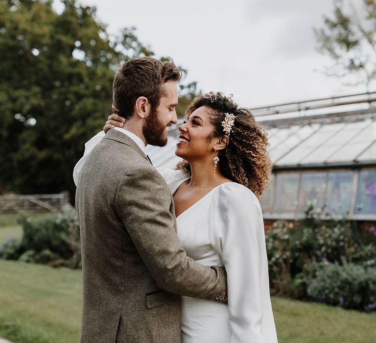 Bride in a satin wedding dress with a front slit looks into the groom's eyes who wears a grey three piece wedding suit