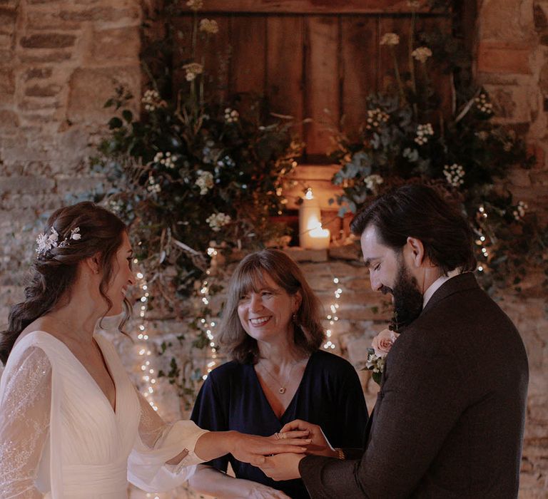 Groom in a brown suit and pale rose buttonhole puts on the bride's wedding ring for their winter wedding in a rustic barn venue 