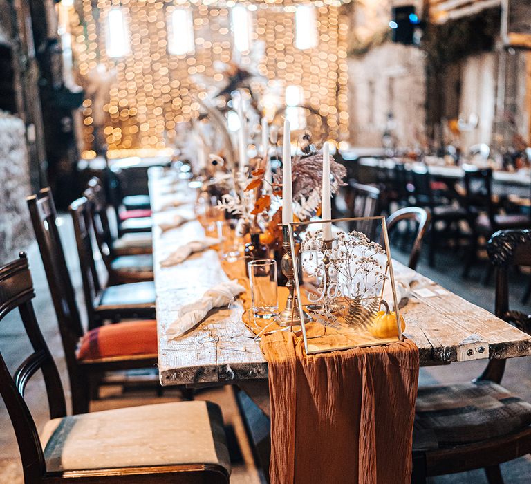 Burnt orange fabric table runner across wooden banquet table complete with white candles and small dried floral centrepieces 