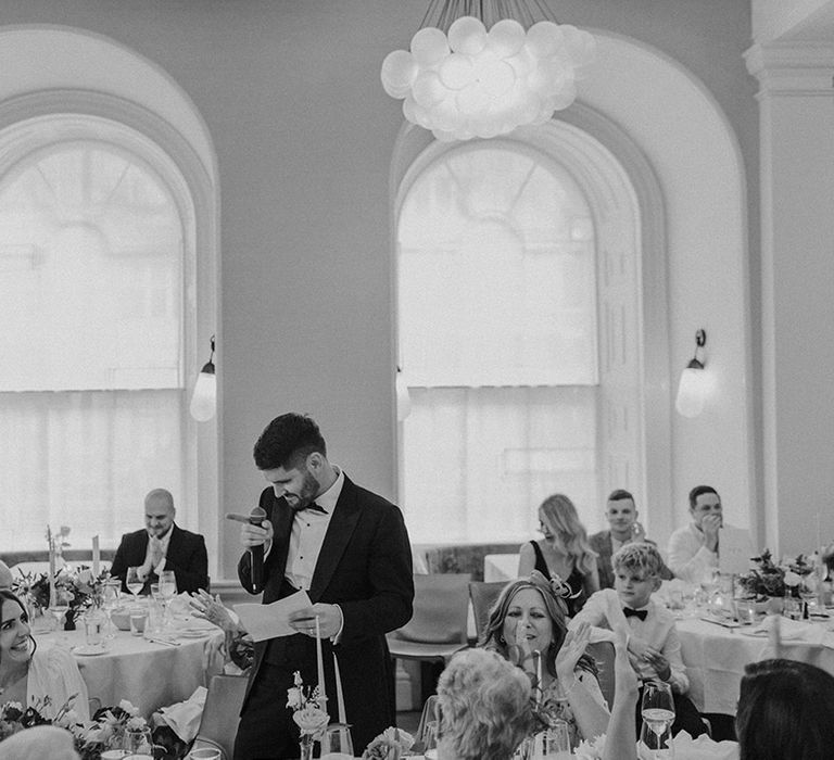 Groom in black tux and bowtie with large boutonniere giving wedding speeches during stylish reception