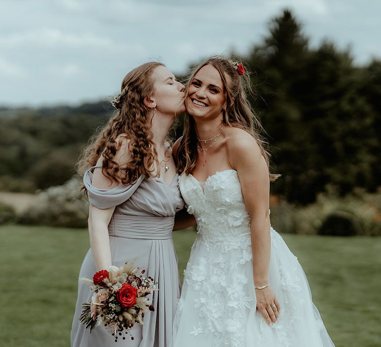 Bridesmaids in a grey off the shoulder dress kisses the bride on the cheek in a strapless flower wedding dress 