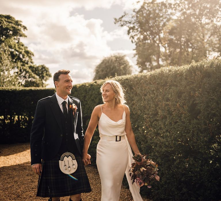 Bride in cowl neck silk slip wedding dress walks alongside her groom in kilt during golden hour