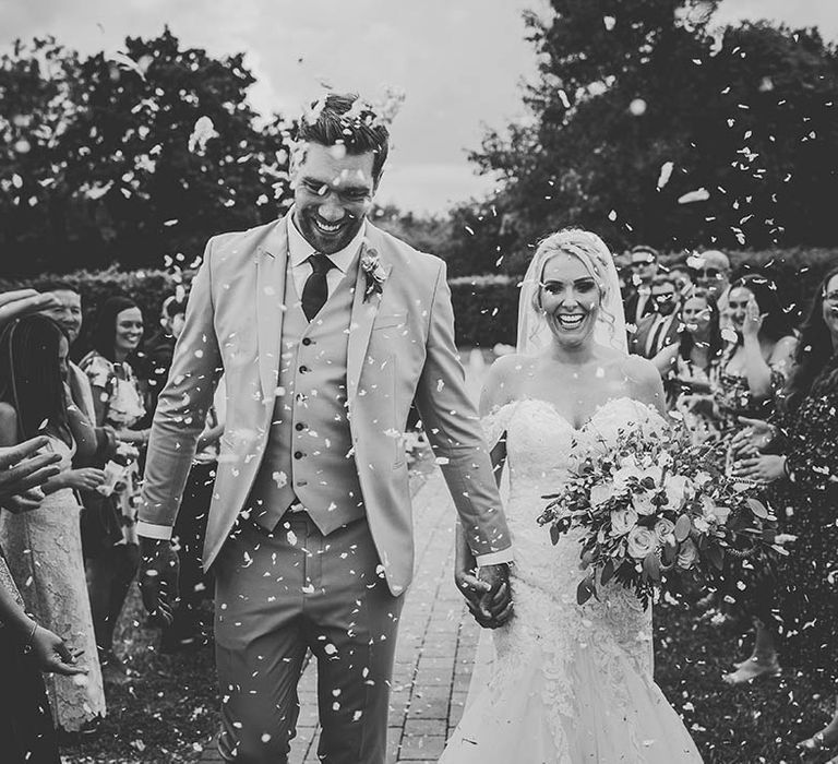 Black and White wedding photography of a confetti shot featuring the Bride wearing a lace wedding dress and the Groom in a three piece suit