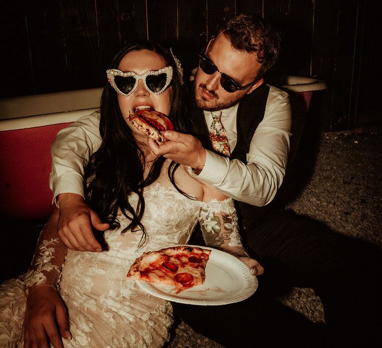  Groom in black waistcoat and flower patterned tie wearing Raybans sunglasses and bride in off the shoulder bridal gown with long, sheer sleeve and lace detailing wearing cat eye bejewelled sunglasses being fed pizza 