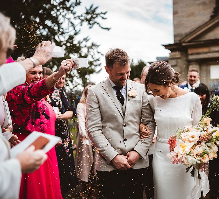 Bride in long satin wedding dress with bridal cover up semi sheer crop top and groom in black suit trousers, grey blazer and light pink rose boutonniere doing confetti walk at The Orangery Ingestre